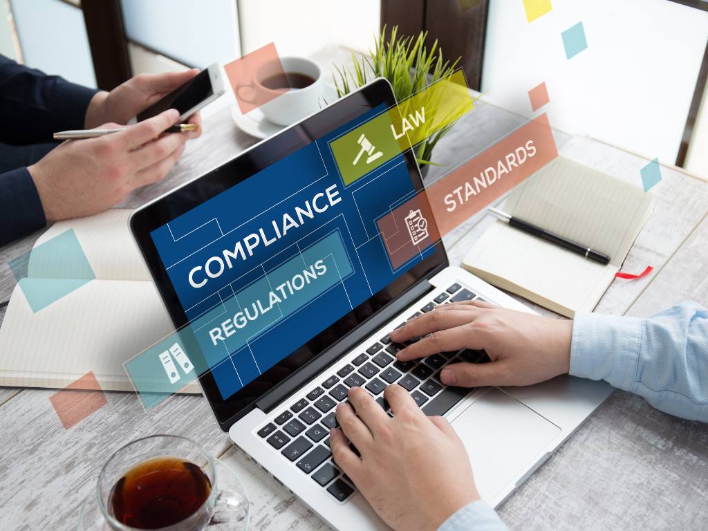 Hands on a laptop keyboard. The words compliance, law, regulations, and standards are superimposed over the screen.