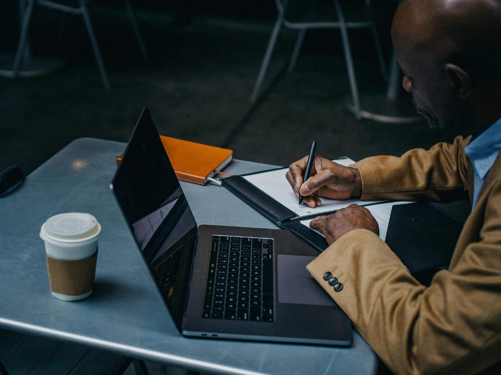 A man sitting at a small table with a laptop and pen and paper.